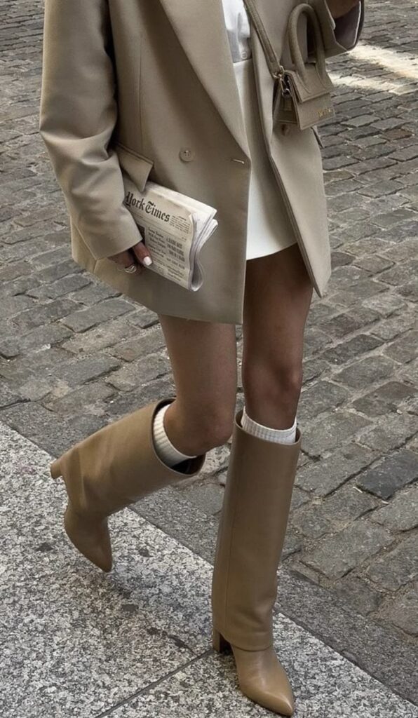Fashionable outfit with beige coat, knee-high boots, mini bag, and a newspaper on a cobblestone street.