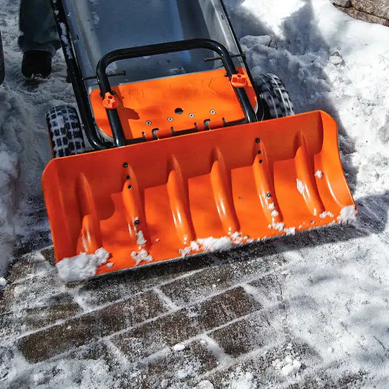 Orange snowblower clearing snow from a brick pathway on a sunny winter day.