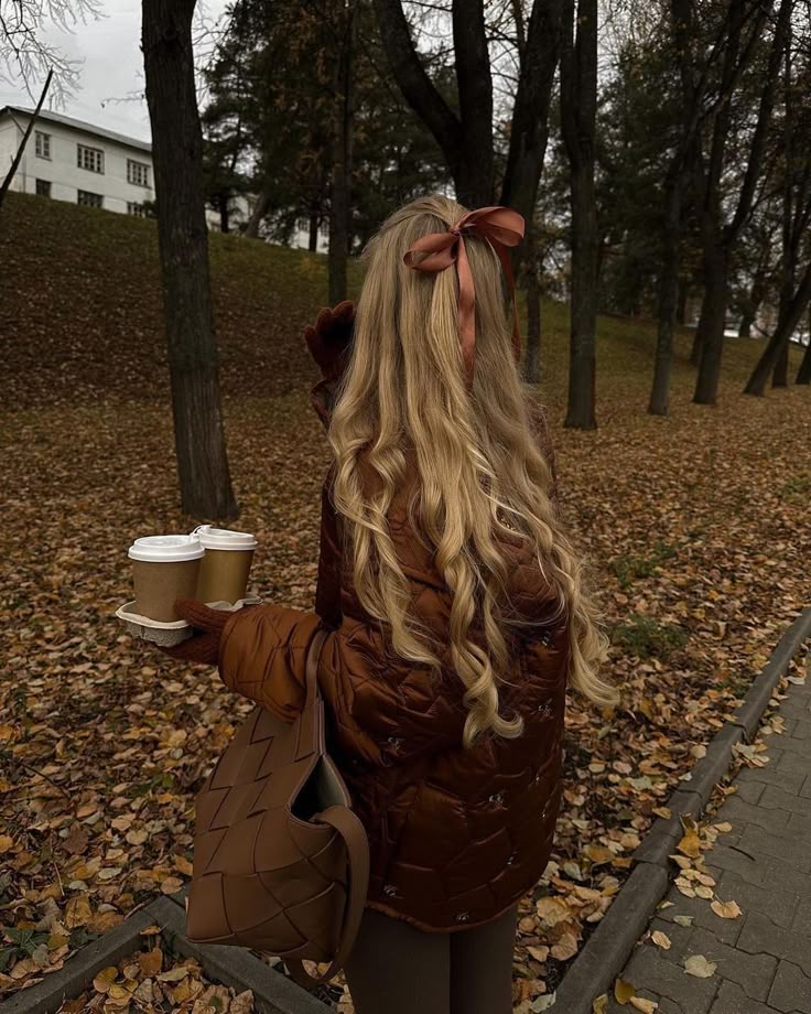 Woman with long blonde hair and bow holding coffee cups in a park with autumn leaves.