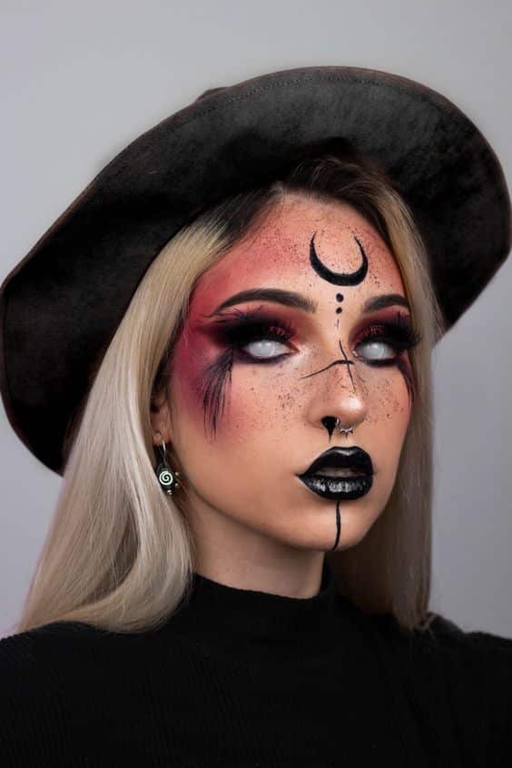 Woman with striking gothic makeup, crescent forehead design, and black hat. Dark lipstick and intense eye makeup complete the look.