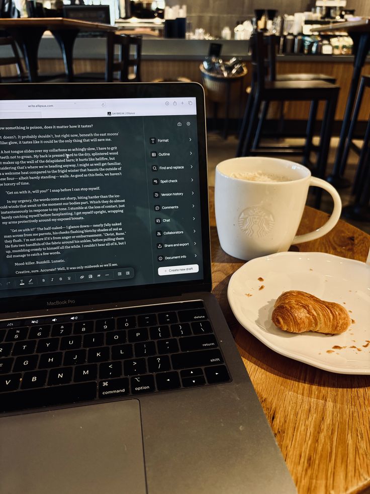 Laptop on table in cafe, open on writing app, with coffee and croissant nearby. Cozy workspace for productivity.