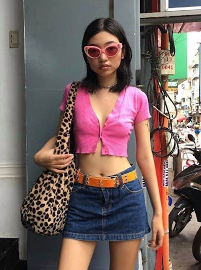 Young woman in pink crop top and denim skirt with leopard print bag and pink sunglasses stands outdoors.