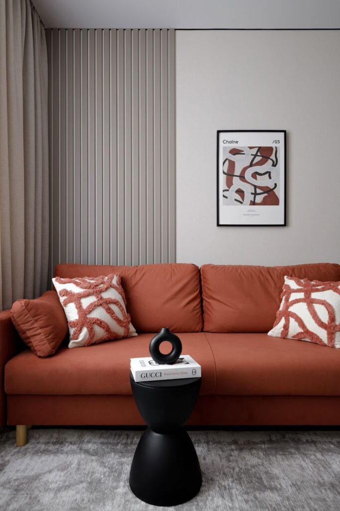 Modern living room with orange sofa, abstract art, and decorative pillows. Minimalist black table holds books.