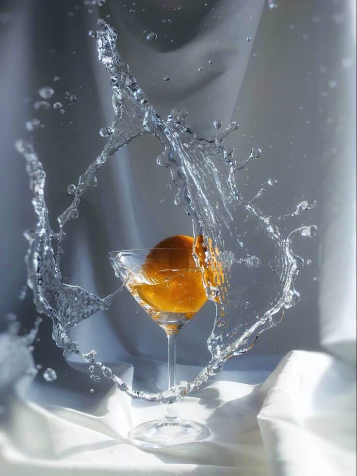 Splashed water surrounds a martini glass with an orange slice, creating an elegant, dynamic scene.