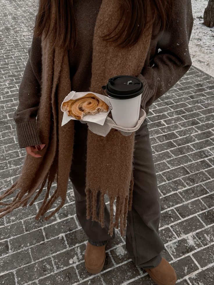 Woman in cozy winter attire holding coffee and pastry on a snowy brick pathway.