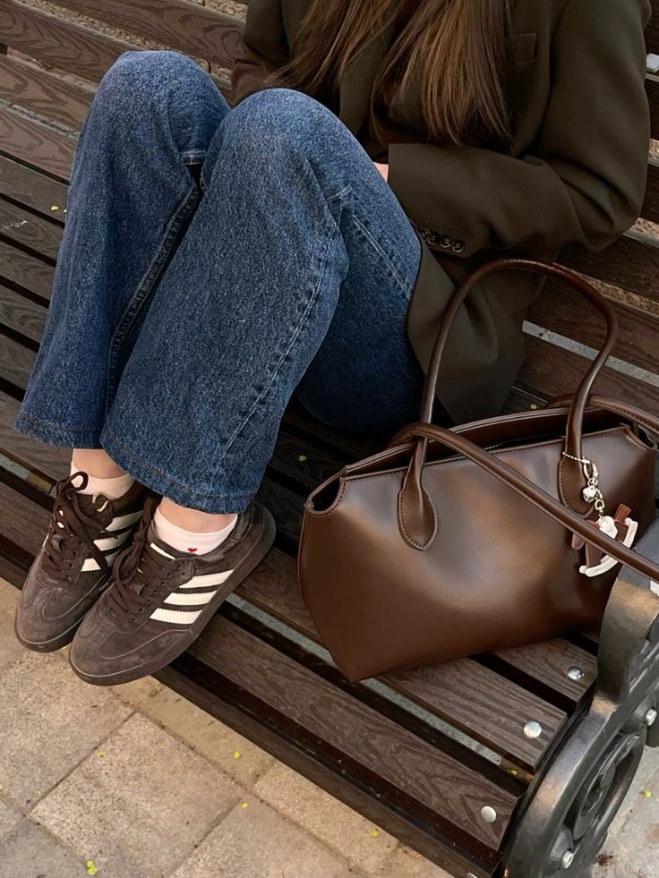 Person sitting on a bench wearing jeans and sneakers, with a brown leather bag beside them.