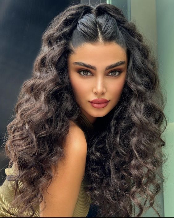 Woman with long, voluminous curly hair and bold makeup, posing indoors against a soft-lit background.