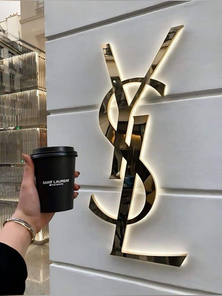 ориброил Hand holding a Saint Laurent coffee cup in front of illuminated YSL logo on a modern building facade. | Sky Rye Design Hand holding a Saint Laurent coffee cup in front of illuminated YSL logo on a modern building facade.