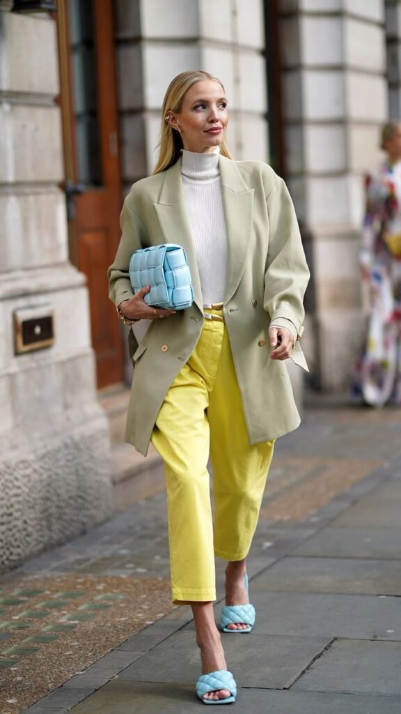 Fashionable woman in pastel blazer and yellow pants, carrying a blue clutch on city street.