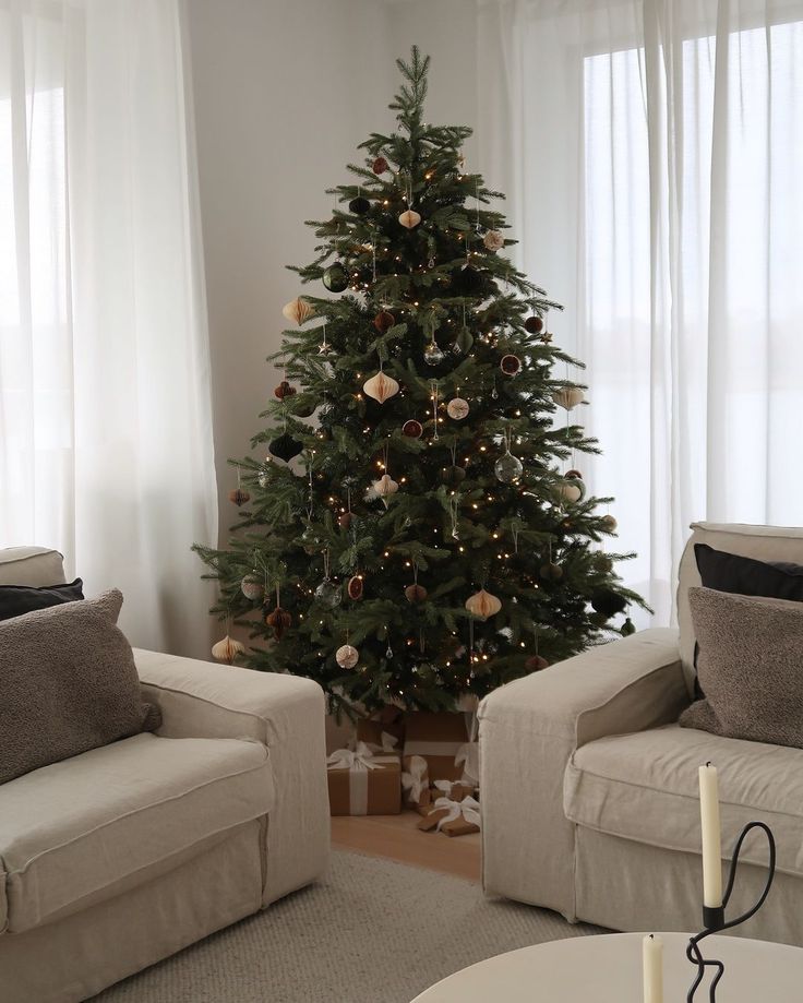 Elegant Christmas tree with ornaments and lights in a cozy living room with beige sofas and large windows.