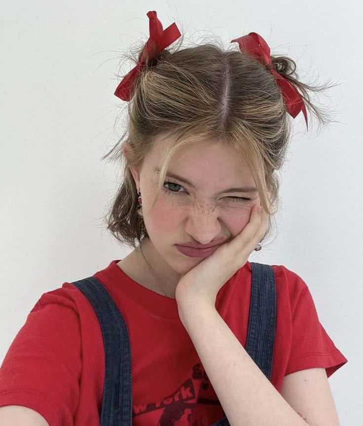 Girl in red shirt with pigtails and red bows, winking with hand on cheek, playful expression.