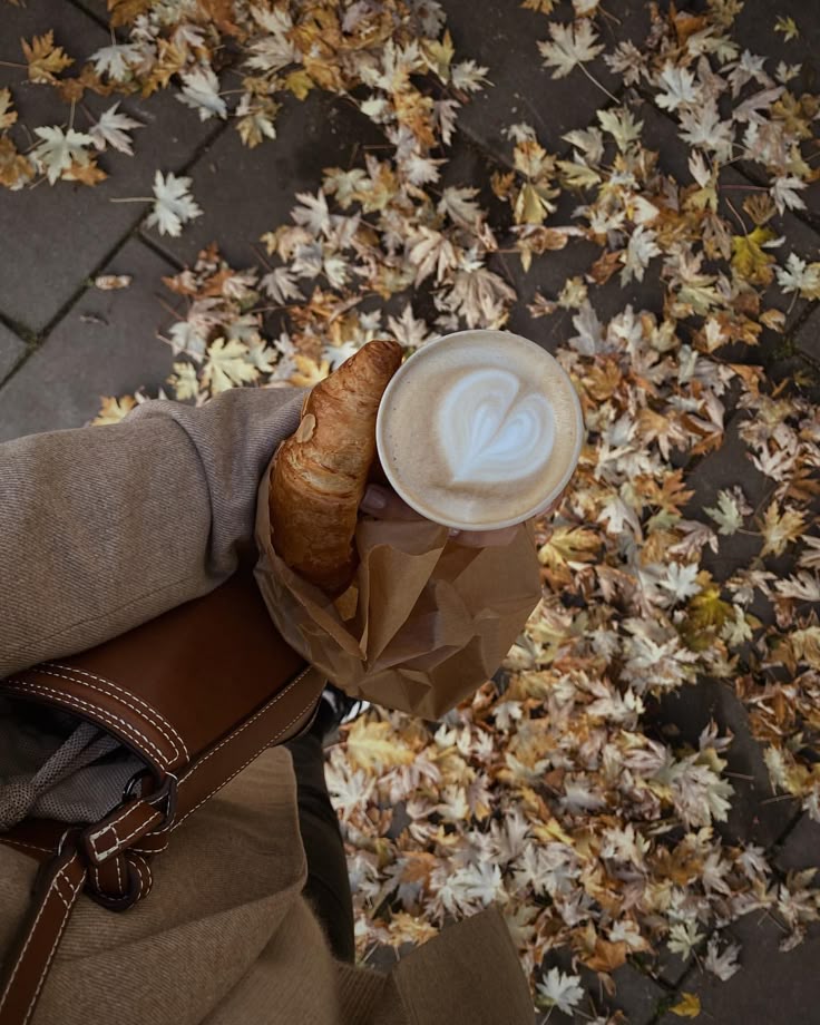 Cafe latte and croissant in hand, autumn leaves covering the ground. Cozy fall vibes on a breezy day.