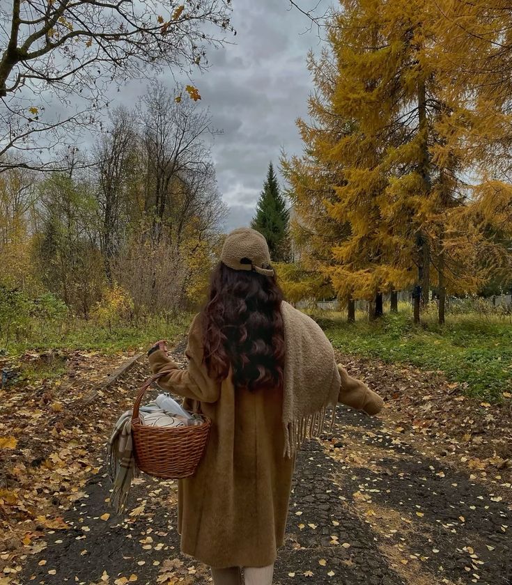 Woman in cozy autumn attire walking down a tree-lined path, carrying a wicker basket under cloudy skies.
