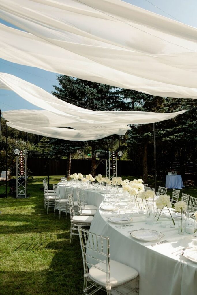 Декор на свадьбе Outdoor elegant event setup with white drapes and decorated tables on a sunny day, ideal for weddings or celebrations. | Sky Rye Design Outdoor elegant event setup with white drapes and decorated tables on a sunny day, ideal for weddings or celebrations.