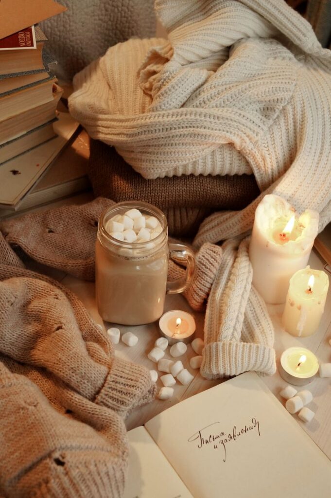 Осенний flatlay Cozy scene with candles, a book, and a mug of hot chocolate and marshmallows, surrounded by warm knitted sweaters. | Sky Rye Design Cozy scene with candles, a book, and a mug of hot chocolate and marshmallows, surrounded by warm knitted sweaters.