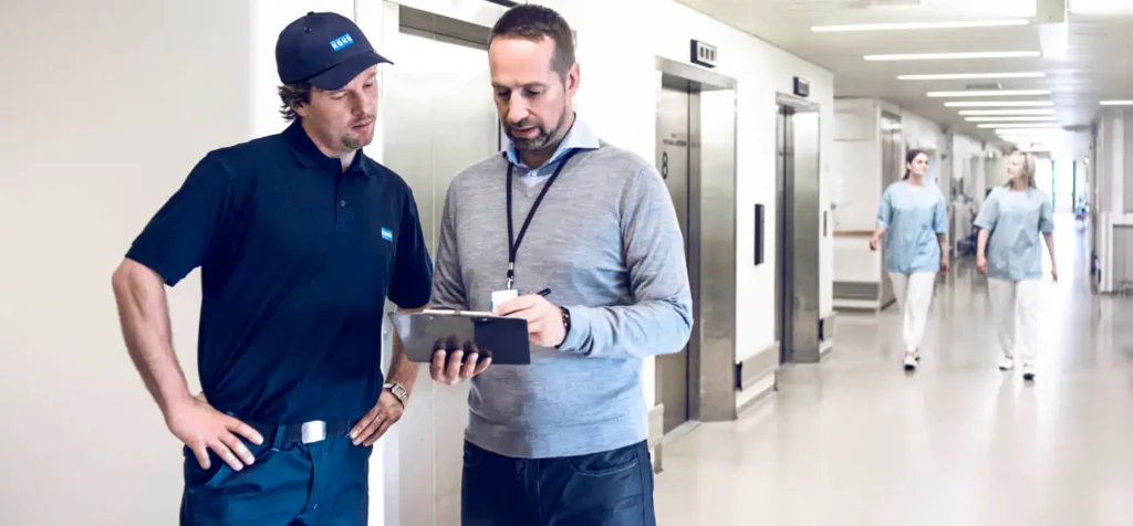 002-kone-medical-1440x670_tcm118-53558 Two men discussing in hallway near elevators, people walk by; professional environment. | Sky Rye Design Two men discussing in hallway near elevators, people walk by; professional environment.