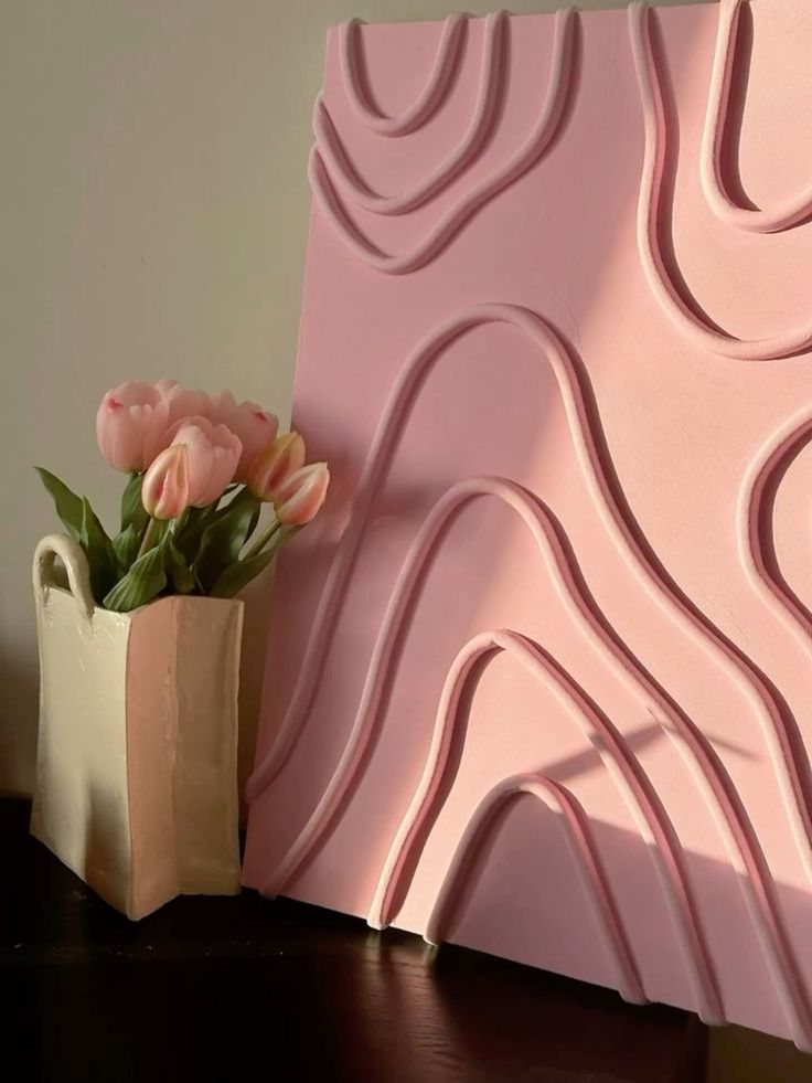 00281925-f1a4-4922-a34d-6c4d78a5cb0f Pink tulips in a beige vase beside pink abstract art with wavy lines, set on a dark table. | Sky Rye Design Pink tulips in a beige vase beside pink abstract art with wavy lines, set on a dark table.