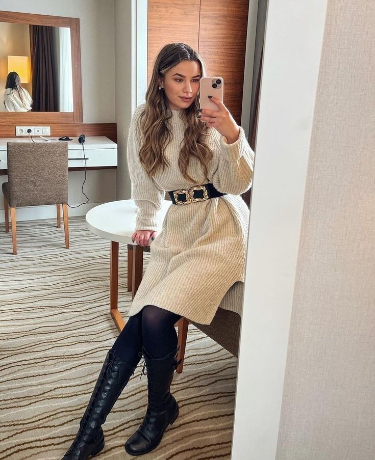 Woman in a cozy beige sweater dress and boots takes a mirror selfie in a modern room.