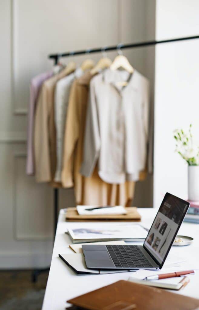 025e3295-3e30-421f-b6d6-5b6c6267d94a Workspace with a laptop on a fashion designer's desk, clothes hanging in the background. | Sky Rye Design Workspace with a laptop on a fashion designer's desk, clothes hanging in the background.