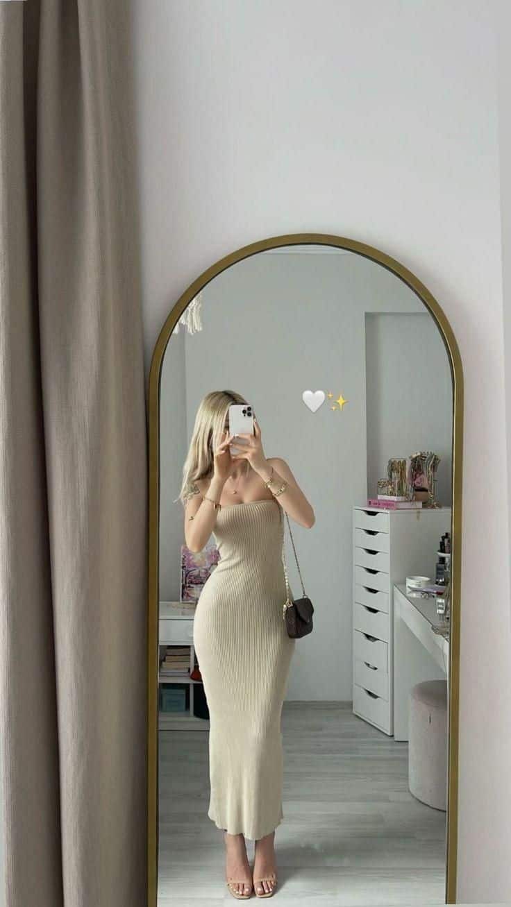Woman takes mirror selfie in beige dress, standing in a modern room with vanity and decor elements.