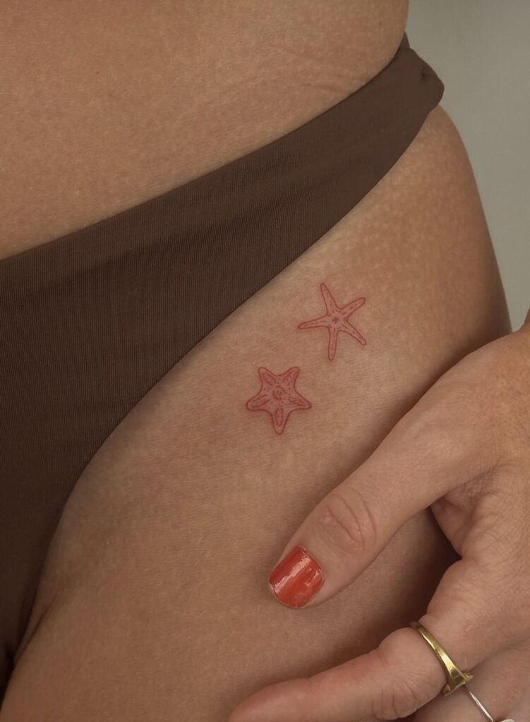07591dd1b9629ba56b669c4a3e932c63 Close-up of two small red starfish tattoos on a person's hip, next to a hand with red painted nails and a gold ring. | Sky Rye Design Close-up of two small red starfish tattoos on a person's hip, next to a hand with red painted nails and a gold ring.