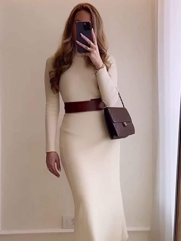 097b51af-3b11-4420-adda-a5edc449784b Woman in beige dress with brown belt, brown boots, and handbag takes a selfie in a minimalist room. | Sky Rye Design Woman in beige dress with brown belt, brown boots, and handbag takes a selfie in a minimalist room.
