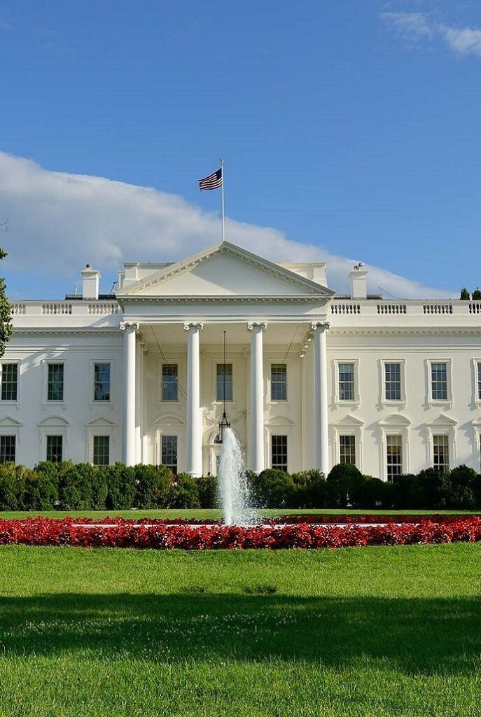 White House with flag, fountain, and lush garden under a clear blue sky.