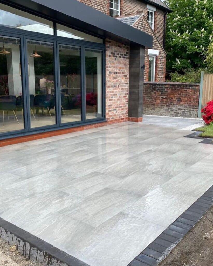 Modern patio with large grey tiles and sliding glass doors leading to a brick house, surrounded by a lush garden.