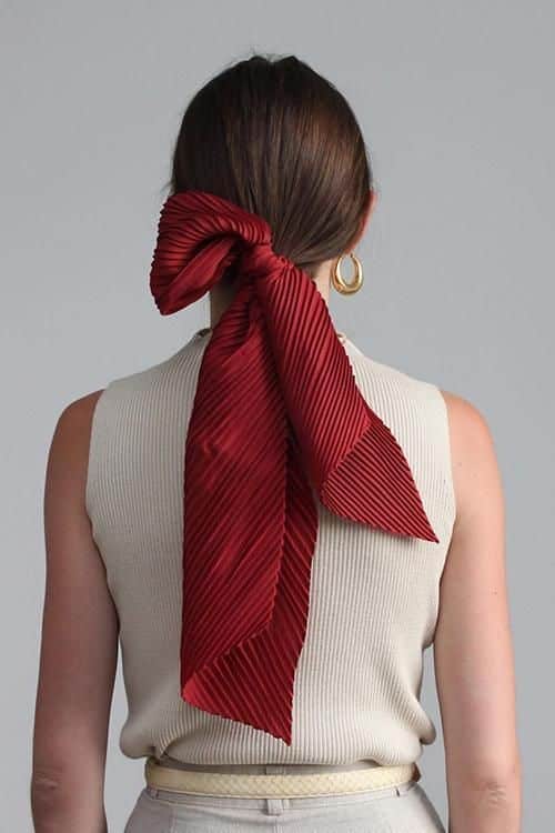 Woman with long red bow in hair, wearing beige sleeveless top and earrings, viewed from behind.