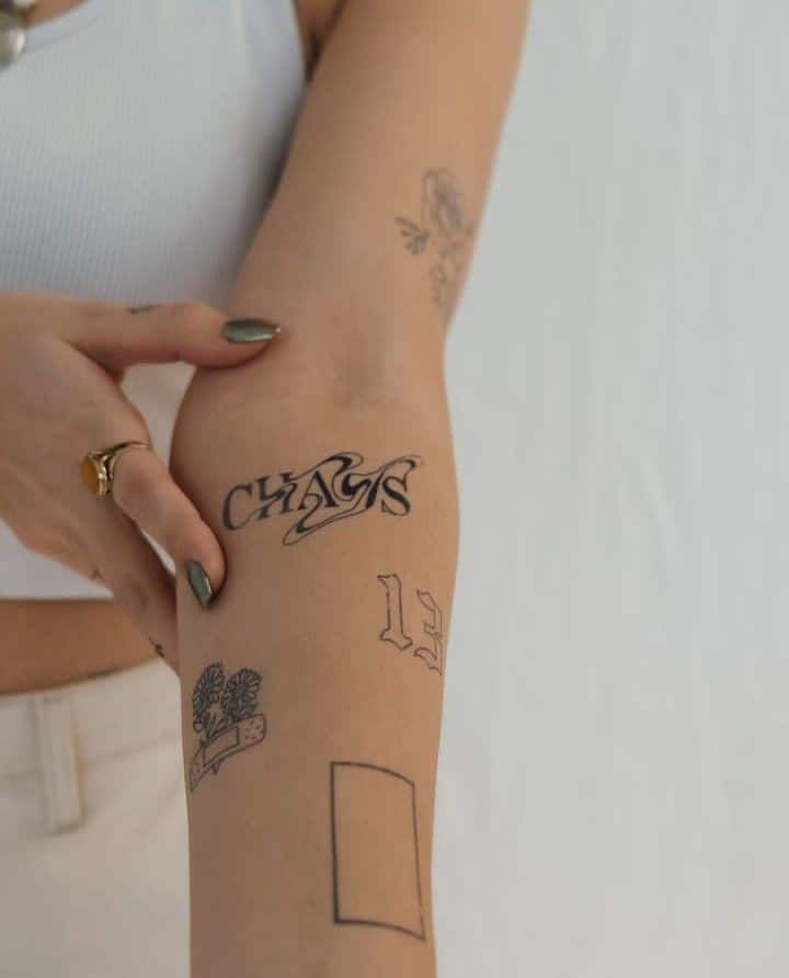 Arm with multiple minimalist tattoos, including CHAOS text and floral designs, on a person wearing a white top.