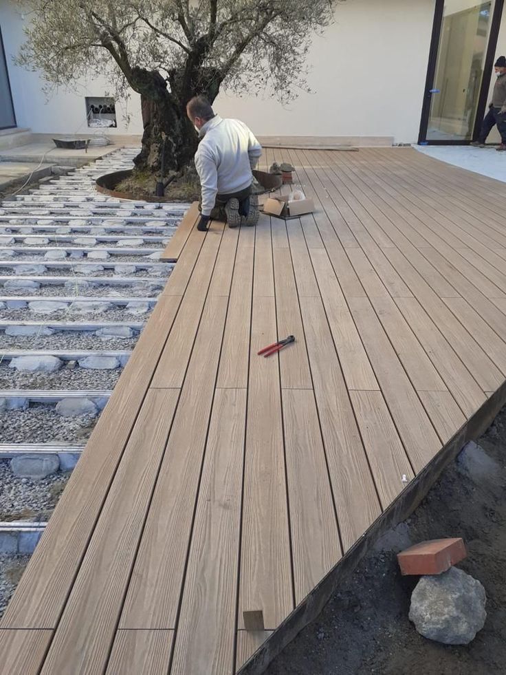 _ 1 Man installing wooden deck planks around an olive tree in a modern backyard. | Sky Rye Design Man installing wooden deck planks around an olive tree in a modern backyard.