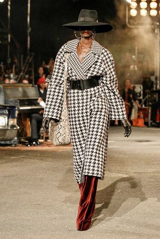 _ 1 Fashion model in houndstooth coat and wide-brim hat on runway, wearing velvet pants and carrying a snakeskin bag. | Sky Rye Design Fashion model in houndstooth coat and wide-brim hat on runway, wearing velvet pants and carrying a snakeskin bag.