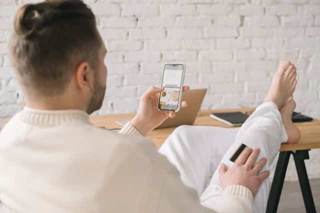 Man shopping online on phone, credit card in hand, sitting relaxed at desk with laptop in bright, modern workspace.
