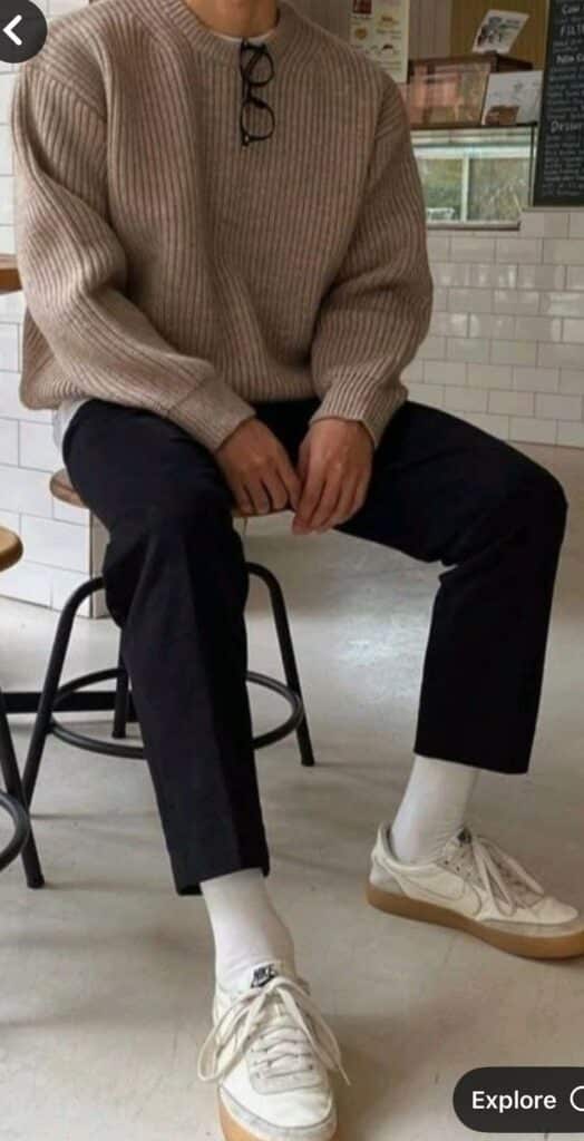 Man in a casual beige sweater and black pants, sitting on a stool, wearing white sneakers and socks.