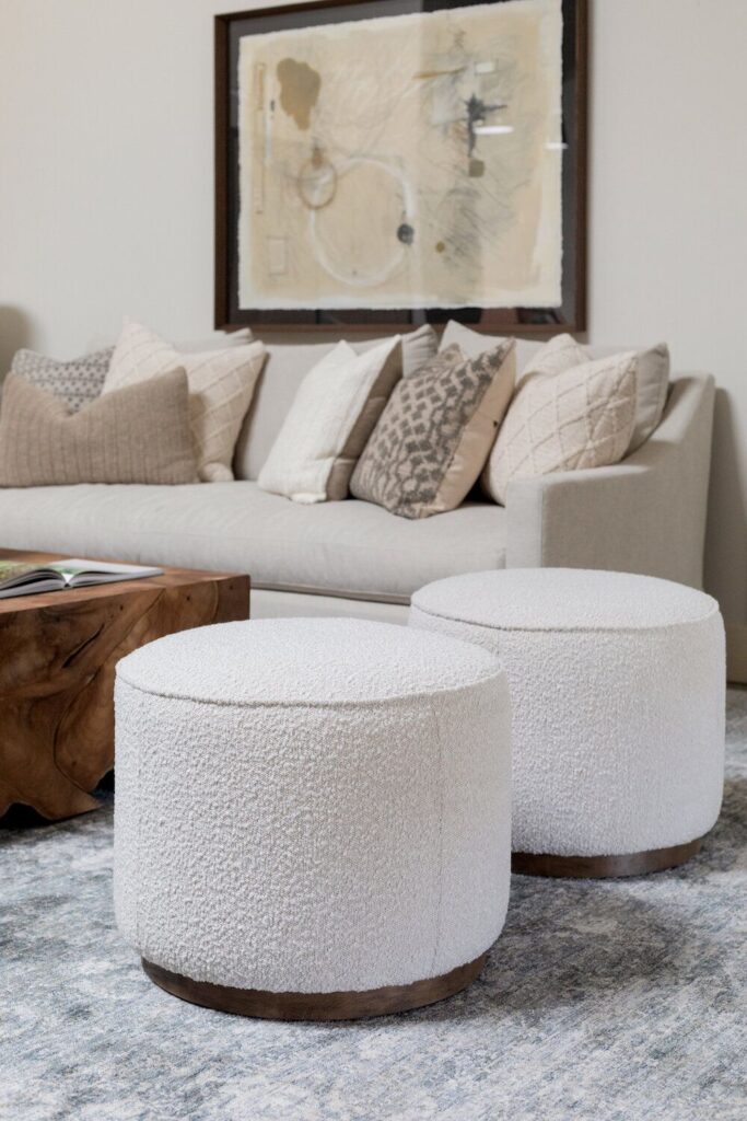 10ea1a4466c240f9e851fc9a46db3070 Modern living room with a beige sofa, patterned pillows, two round ottomans, and abstract wall art. | Sky Rye Design Modern living room with a beige sofa, patterned pillows, two round ottomans, and abstract wall art.