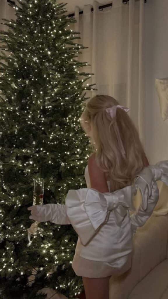 Woman in elegant dress by a lit Christmas tree holding a champagne glass, creating a festive holiday scene.