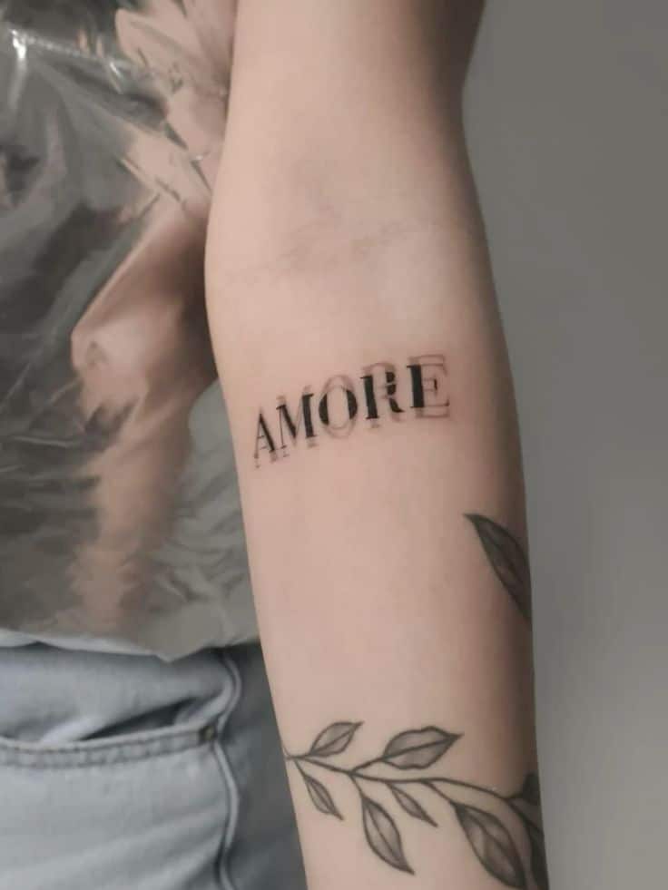 Arm tattoo of the word AMORE in bold black font with botanical design on forearm.