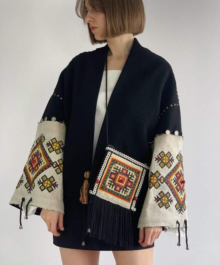 Woman in embroidered jacket with geometric patterns and fringe standing against a plain background.