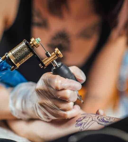 _ 12 Tattoo artist using a machine to ink a floral design on a person's arm in a studio. | Sky Rye Design Tattoo artist using a machine to ink a floral design on a person's arm in a studio.