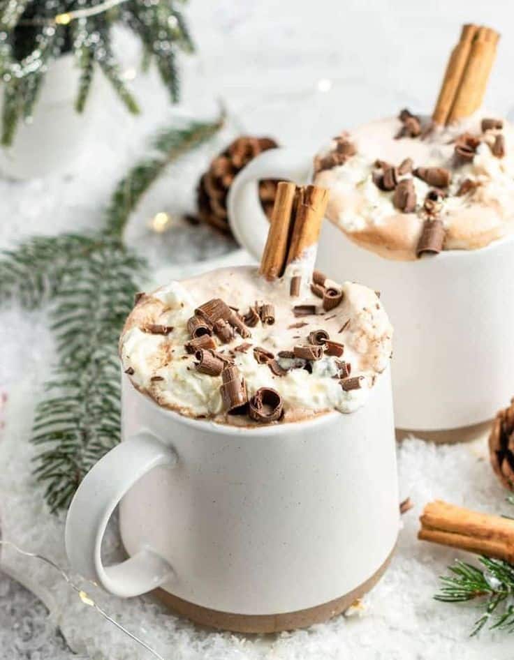 Creamy hot chocolate with whipped cream, chocolate shavings, and cinnamon sticks in white mugs, snowy setting background.