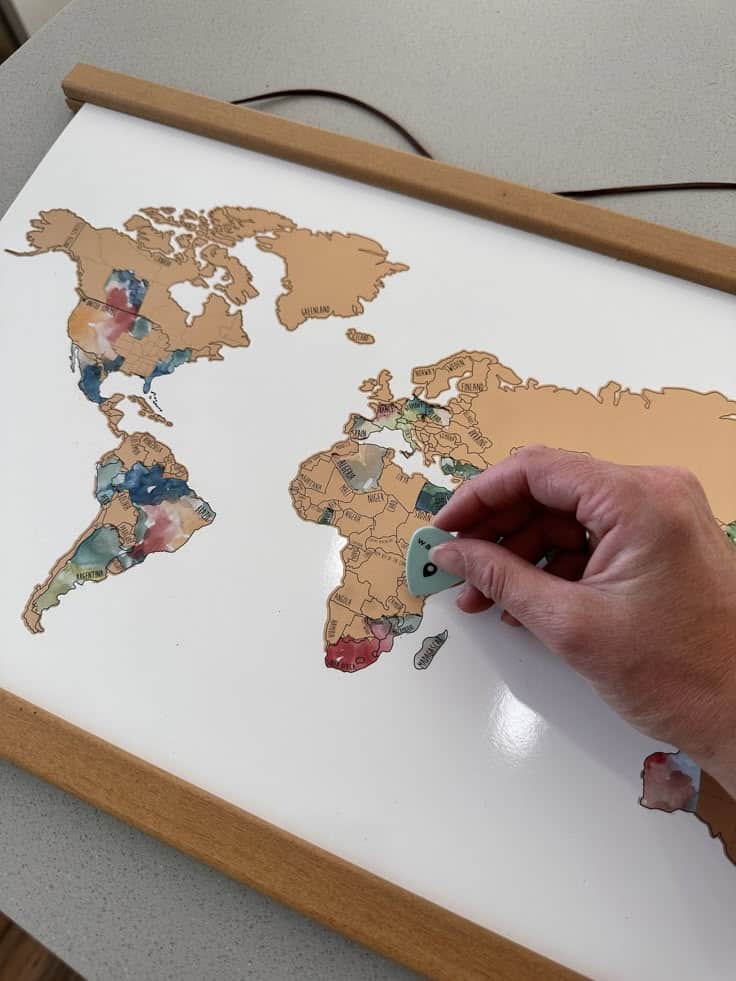 Hand scraping world map poster using a guitar pick to reveal colorful countries beneath brown coating.