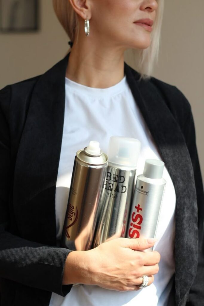 Woman holding hair styling products, including styling spray and finishing products, wearing a black blazer.