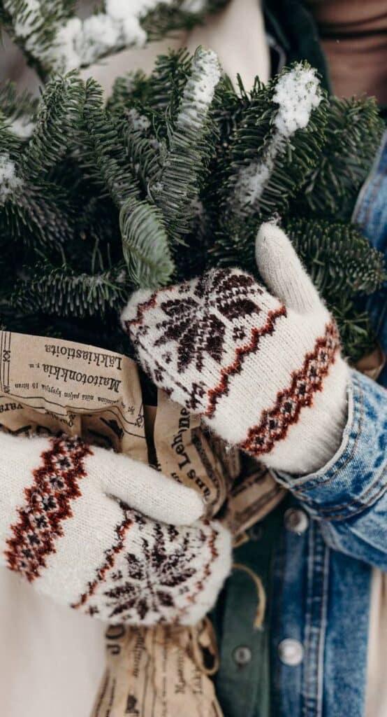 _ 13 Cozy knit mittens holding a snow-dusted pine branch wrapped in newspaper, embodying winter warmth and holiday spirit. | Sky Rye Design Cozy knit mittens holding a snow-dusted pine branch wrapped in newspaper, embodying winter warmth and holiday spirit.