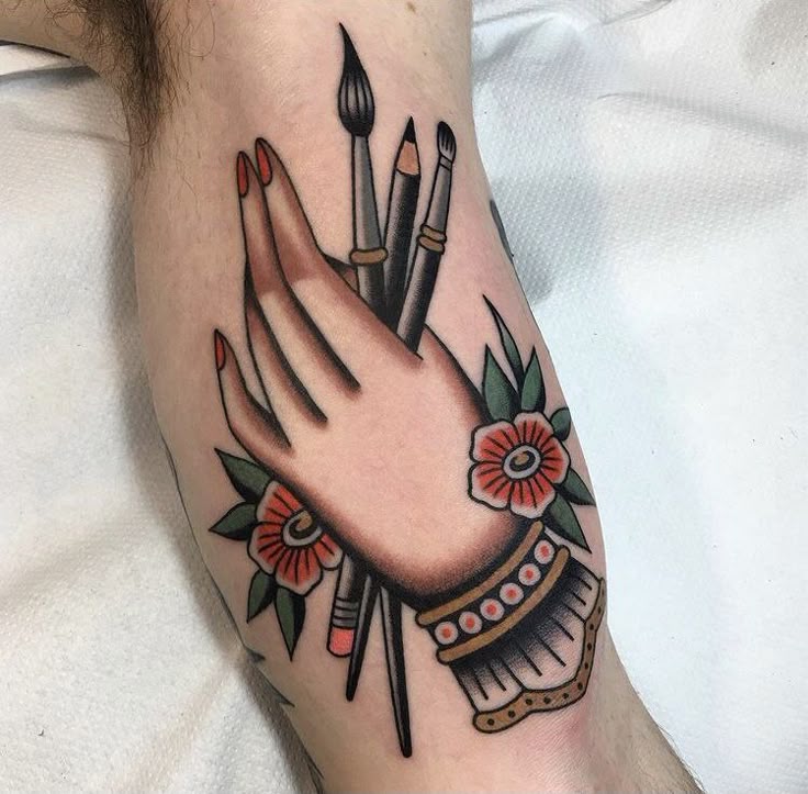 Tattoo of a hand holding art brushes, surrounded by flowers, on a person's arm.