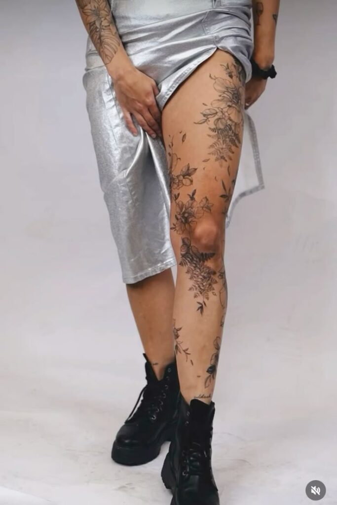 _ 13 Person displays floral leg tattoos in a silver skirt and black boots, showcasing body art fashion. | Sky Rye Design Person displays floral leg tattoos in a silver skirt and black boots, showcasing body art fashion.
