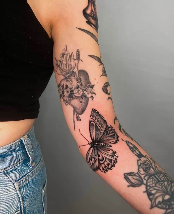 Arm with detailed tattoos: butterfly, heart with flowers and dagger, black tank top, and denim jeans.