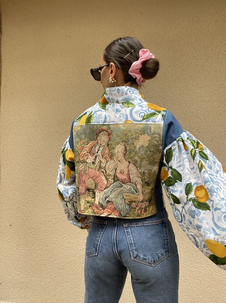 Woman in colorful tapestry jacket and jeans, hair in bun with pink scrunchie, facing beige wall.
