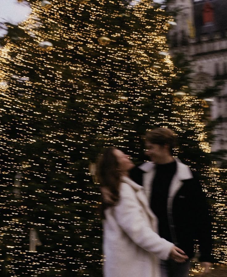 Couple smiling joyfully in front of a large Christmas tree adorned with sparkling lights, creating a festive atmosphere.