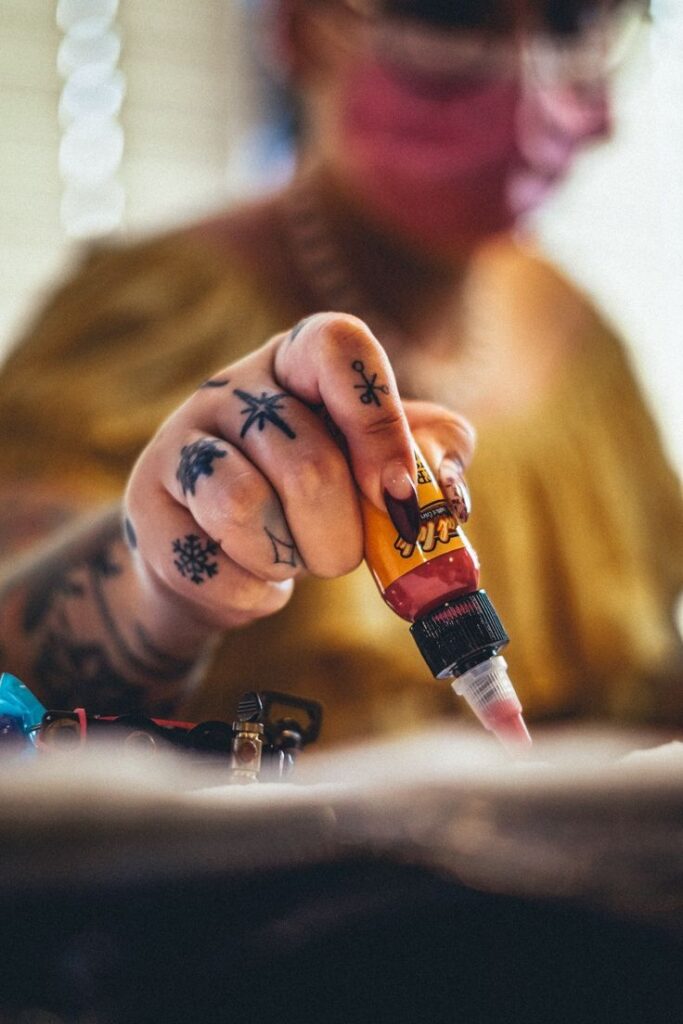 _ 14 Tattoo artist filling a needle with ink, showcasing tattooed hand for precision craft creation. | Sky Rye Design Tattoo artist filling a needle with ink, showcasing tattooed hand for precision craft creation.