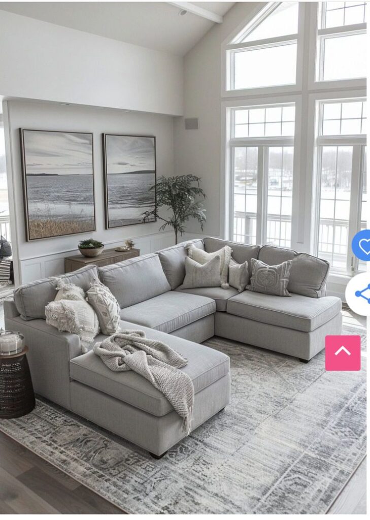 Modern living room with gray sectional sofa, large windows, coastal art, and cozy decor on patterned rug.